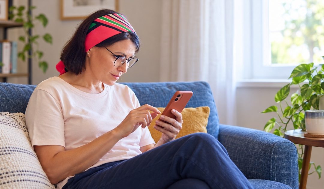 A woman with glasses and a striped headband reads on her smartphone while seated on a blue sofa.